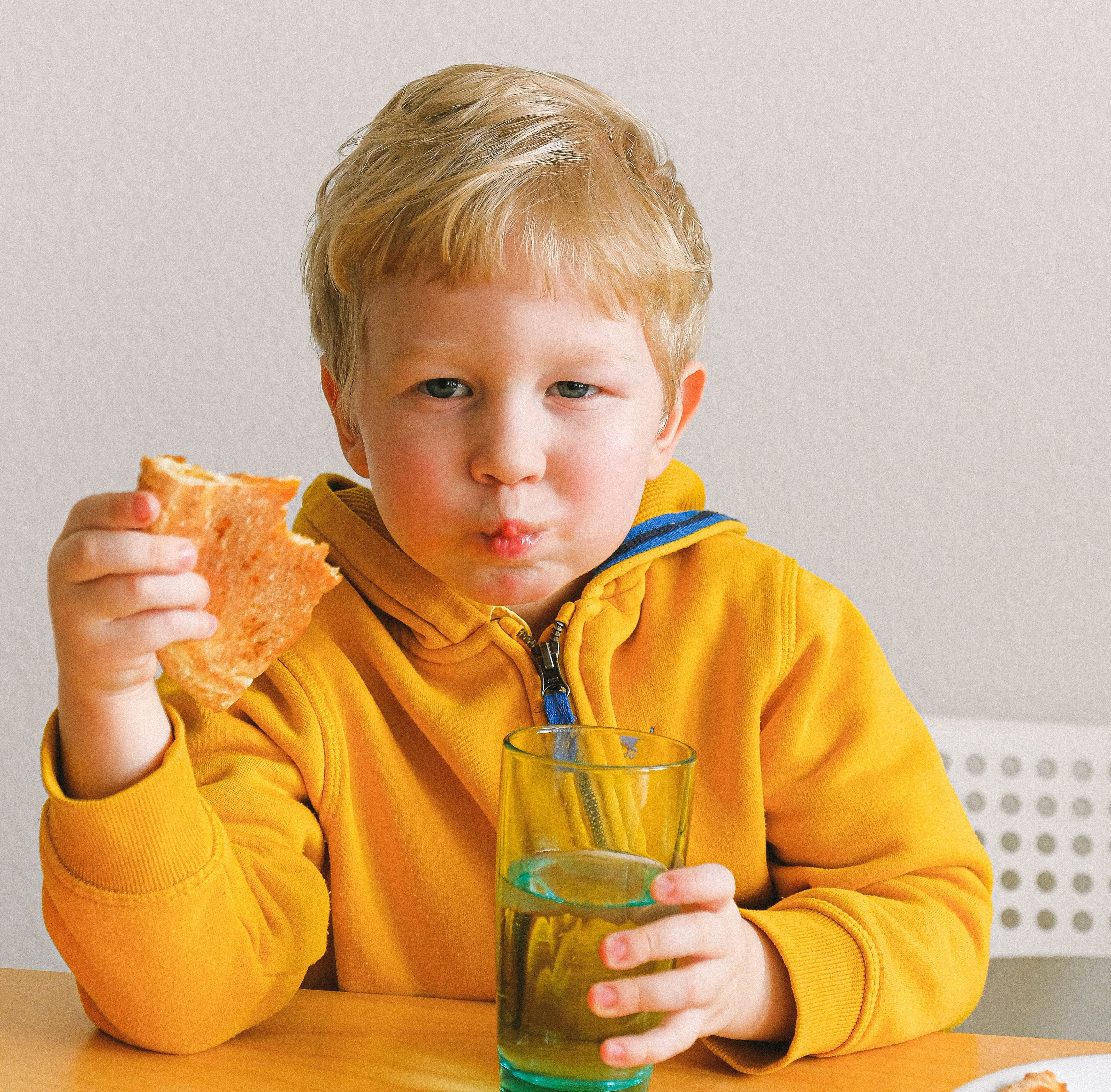 A blonde boy in a yellow fleece jacket eats a slice of pizza and clasps a glass of water.