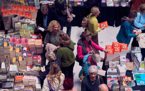 An overhead shot of a large group of people milling between tables stacked high with books. People browse books, chat to one another, and make purchases.