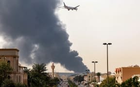 An Emirates aircraft prepares for landing as a smoke plume rises from an ongoing fire near Dubai International Airport in Dubai on March 16, 2026. Flights were gradually resuming at Dubai airport on March 16, previously the world's busiest for international flights, the airport operator said, after a "drone-related incident" sparked a fuel tank fire nearby, as Iran kept up its Gulf attacks.
