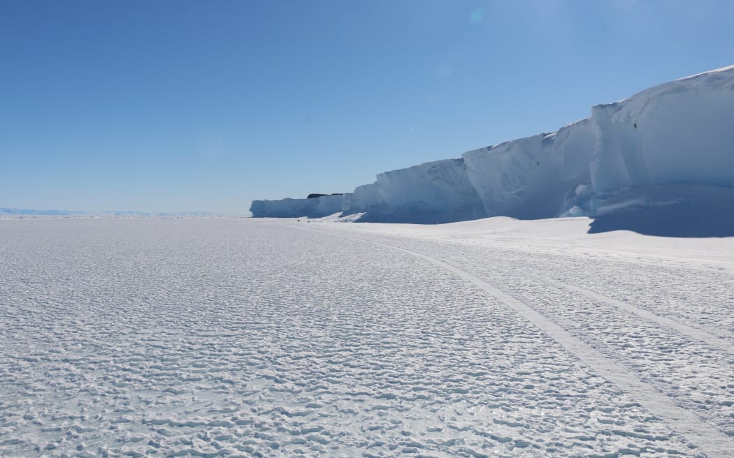 A wide shot of a flat expanse of ice with one set of vehicle tracks alongside a cliff of ice on the right. The sky is blue and cloudless.