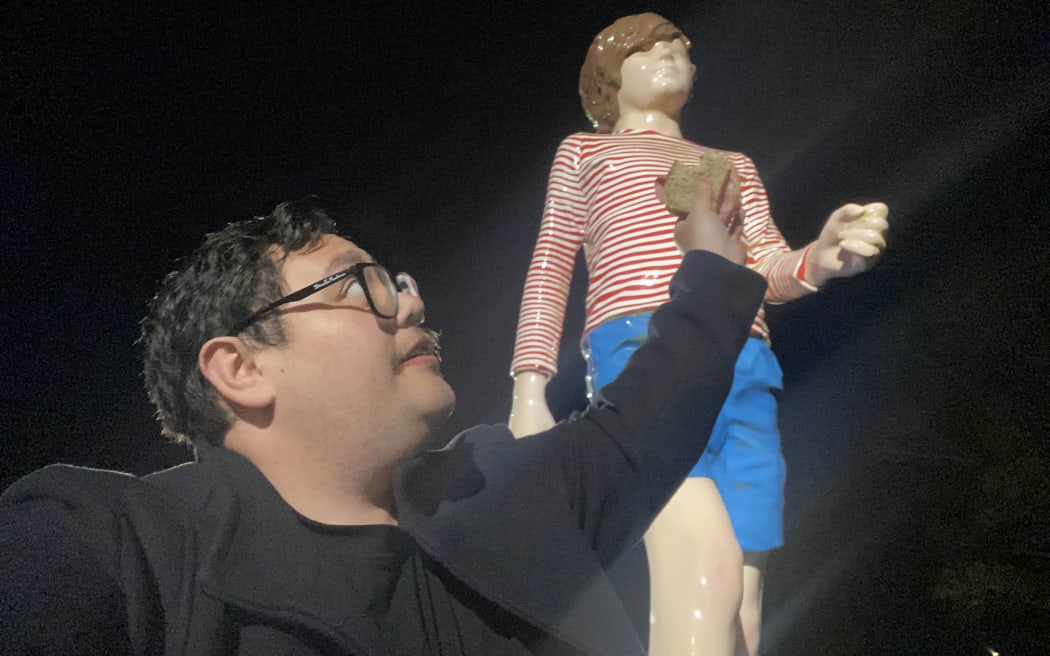 A man in a coat holds up a freshly cooked bacon sandwich up to a giant statue of a kid in blue shorts and a red and white striped shirt.