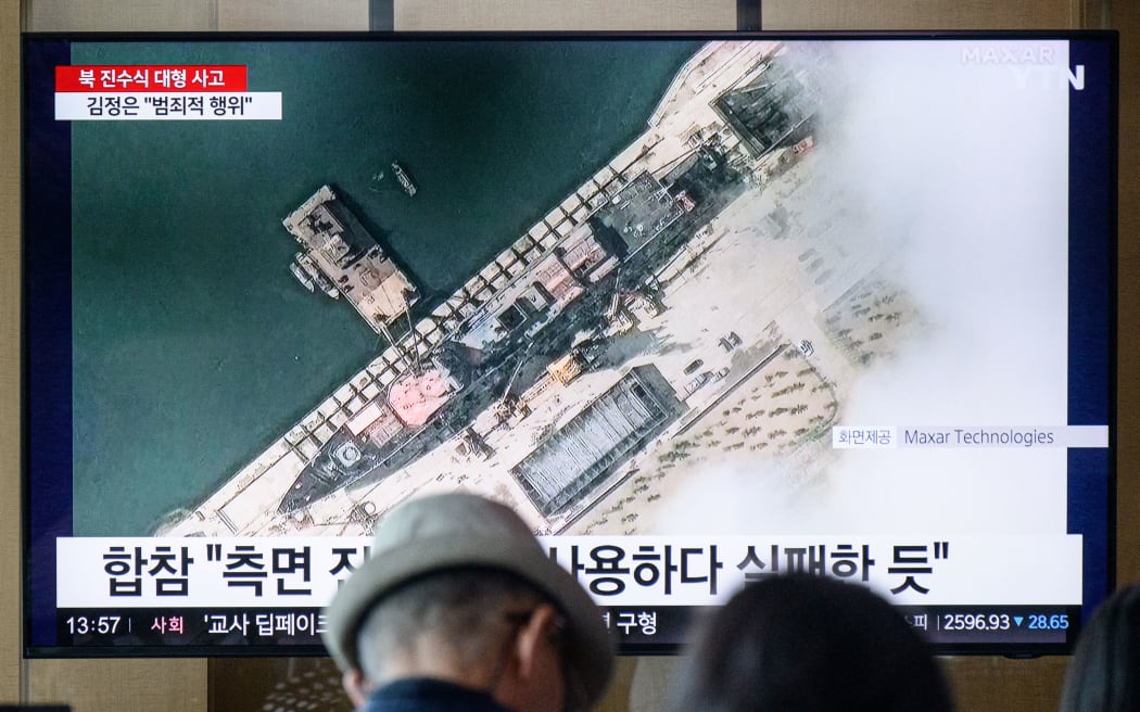 In a photo taken in Seoul on May 22, 2025, people sit near a television showing news footage with a satellite still image courtesy of Maxar Technologies showing a general view of a new North Korean warship at the harbour ahead of its launch in Chongjin. A major accident occurred at the launch ceremony for a new North Korean naval destroyer, state media reported on May 22, with leader Kim Jong Un calling the mishap a "criminal act". (Photo by ANTHONY WALLACE / AFP)