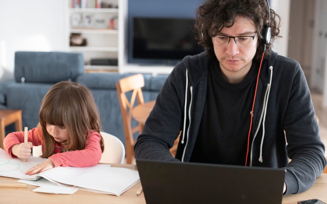 Young father working from home with little daughter drawing during covid-19 lockdown. Child drawing next to dad who is smartworking with laptop and headphones for social isolation. Front view.