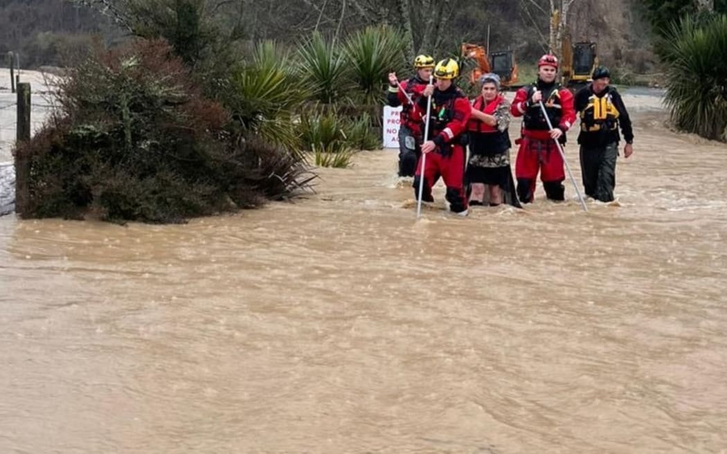 Two people are guided across dangerous floodwaters in Tasman on Friday 11 July, 2025, by members from Fire and Emergency NZ's specialist water response teams from Christchurch and Nelson, using long poles to test what lies under the water.