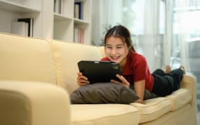 Happy young woman enjoying content on her digital tablet, lying on a sofa at home.