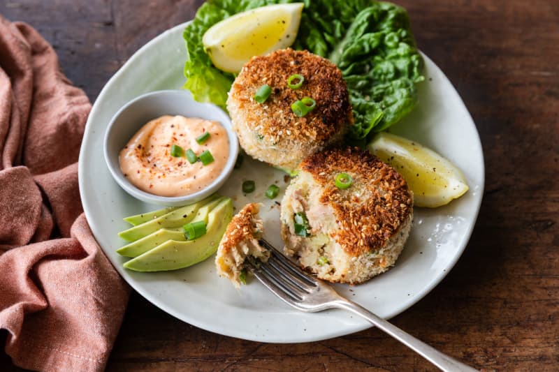A plate containing two green curry potato and tuna fish cakes, accompanied with sliced avocado, lemon wedges and lettuce.