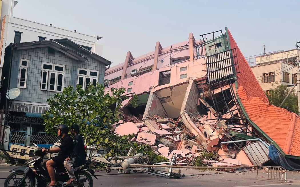 People drive on a motorbike past a collapsed building in Mandalay on March 28, 2025, after an earthquake in central Myanmar. A powerful earthquake rocked central Myanmar on March 28, buckling roads in capital Naypyidaw, damaging buildings and forcing people to flee into the streets in neighbouring Thailand.