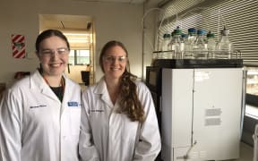 Two women in white lab coats and lab goggles stand in a lab next to a large machine with about a dozen glass bottles on top of it. The glass bottles are half-filled with clear liquid and have tubes running from them into the machine.