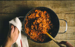 A black cast iron pot containing a cooked lentil stew on a wooden board is being stirred with a wooden spoon while one hand holds the handle with a white cloth.