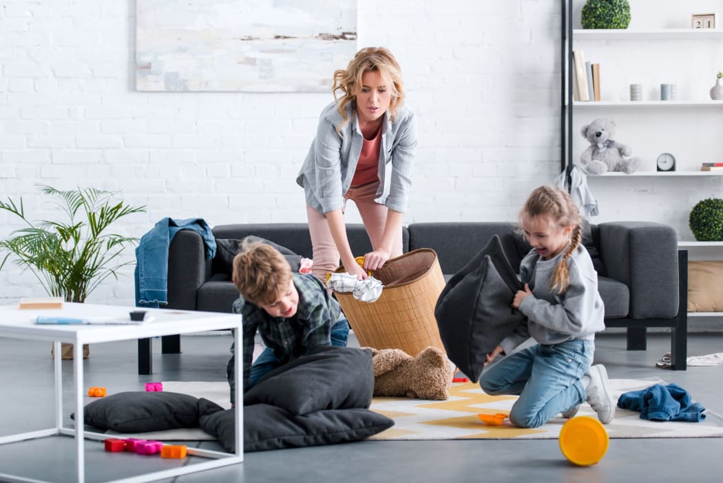 A photo of an emotional tired mother putting toys in basket while naughty kids fighting with pillows