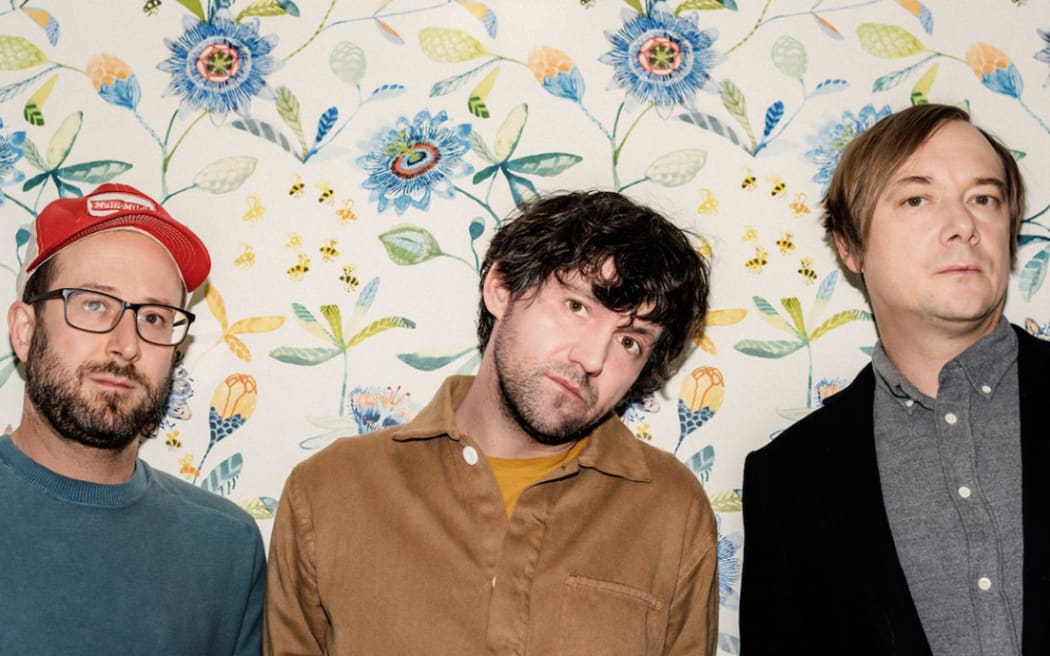 The three members of Bright Eyes lined up against a flowery wall