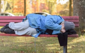 Homeless elderly woman sleeping rough in a park curled up against the cold autumn weather on a rustic wooden bench