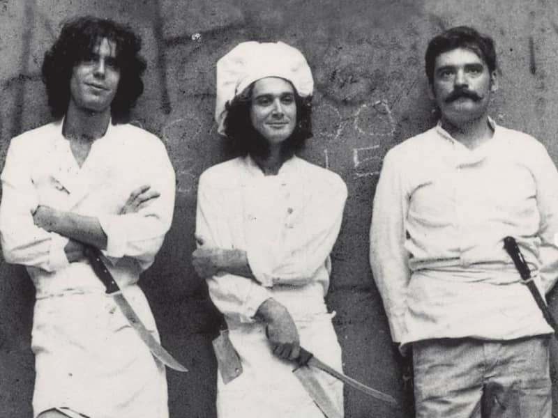 Three men wearing white aprons and carrying knifes are lined up against a wall in a black and white photo.