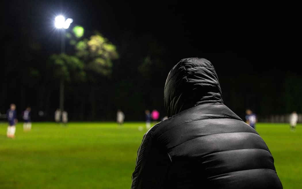A figure in a hooded jacket watches from the sidelines during an amatuer competition football game in Auckland one weekday evening.