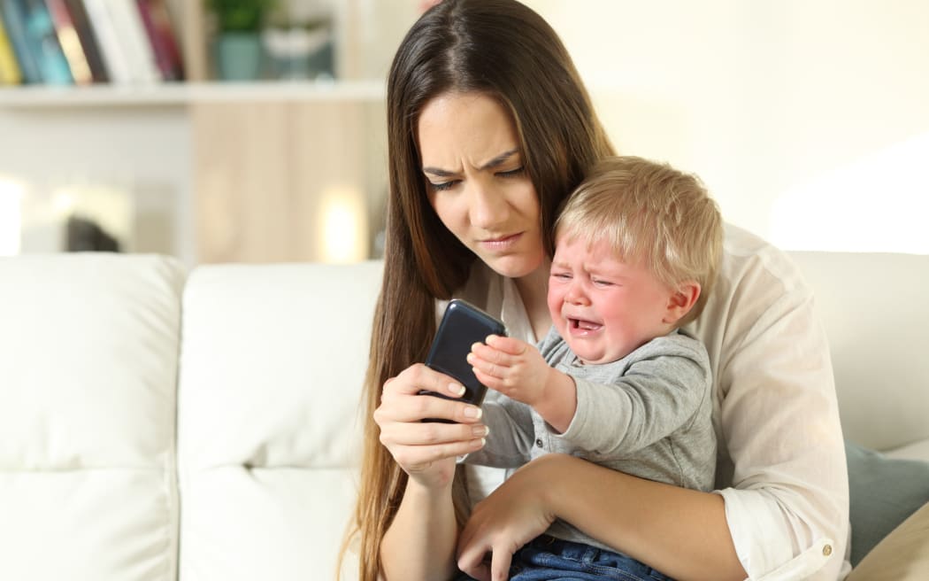 A photo of a baby having a tantrum and fighting with his mother for a smart phone sitting on a couch in the living room at home