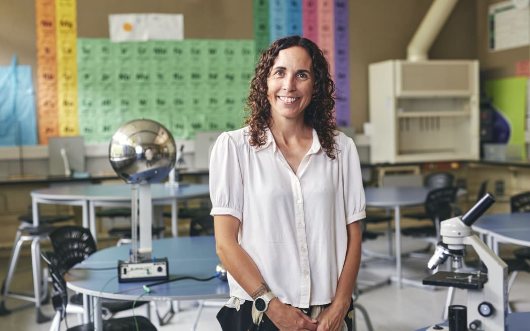 A woman with long curly brown hair stands in a school science lab with a huge periodic table behind her, and a microscope to her left. She is wearing a short-sleeved white button-up shirt and looks happy.