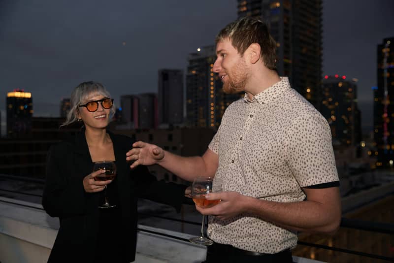 Two people holding wine glasses, standing on a building rooftop in a city.