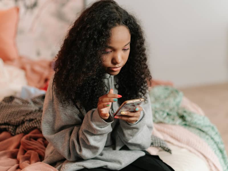 A young girl with curly hair wears a grey sweater and looks at her phone.