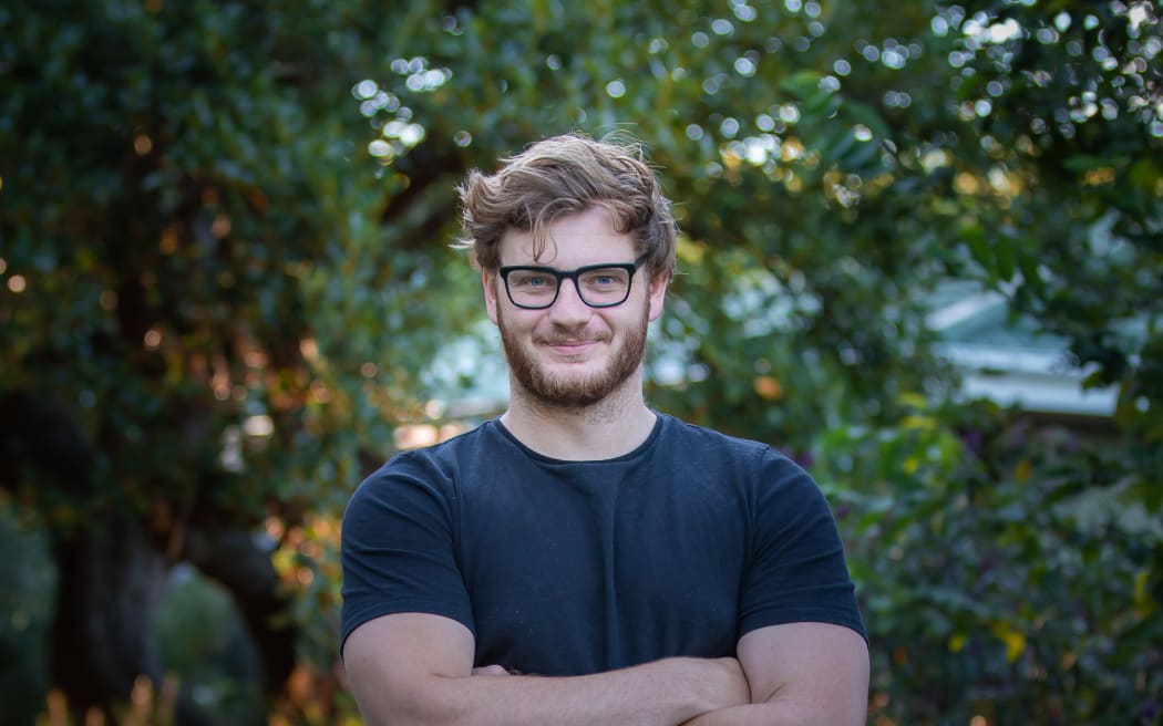 Ryan smiles at the camera. He stands with his arms folded in a garden with leafy trees all around him. He wears glasses and a black tshirt.