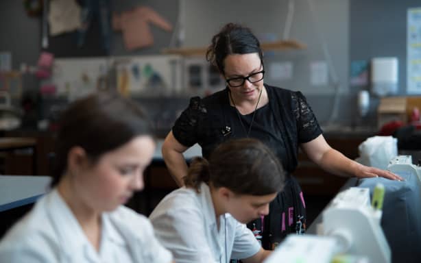 A classroom image of Nan teaching. She is in focus, towards the back of the picture, with her hand on a sewing machine and her head leaning towards a student. The two students towards the foreground are slightly blurry and leaning over two other sewing machines.