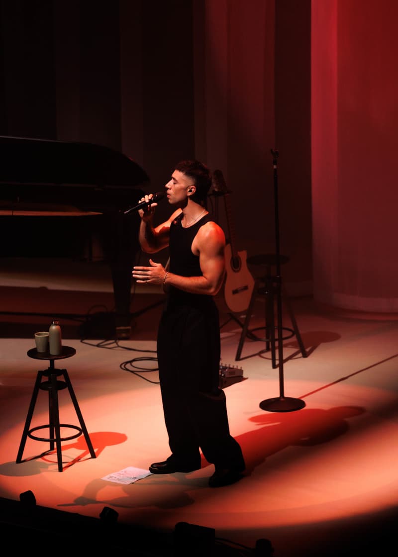 TEEKS, a muscular man in a black singlet and black jeans, sings into a microhpne with members of an orchestra and a red velvet curtain behind him.