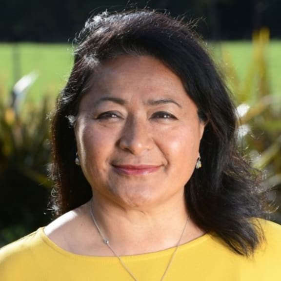 Mena smiles at the camera. She is outdoors standing on a lawn, wearing a yellow top.
