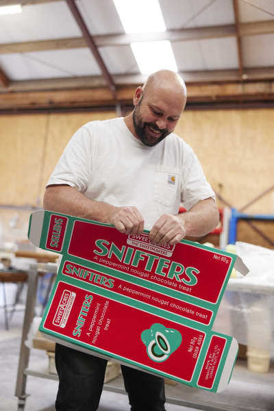 A bald bearded man in a white t-shirt holds a green and red box that says the word 'Snifters'