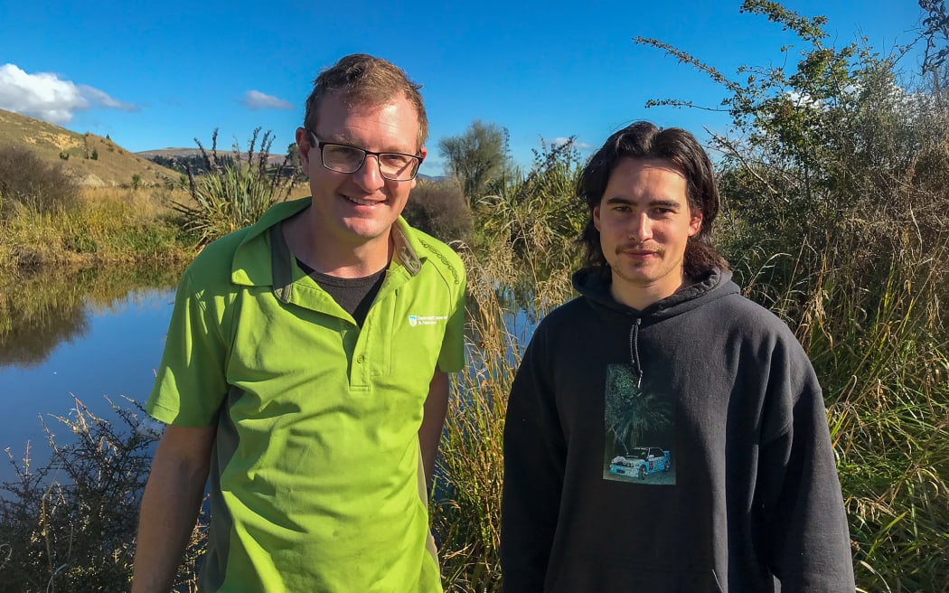 Two people stand in front of a wetland pool surrounded by vegetation. Both are smiling at the camera. The man on the left has glasses and a bright green collared t-shirt. The man on the right is wearing a dark coloured hoodie. The sky is blue and cloudless.