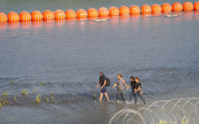 (FILES) Migrants walk between Concertina wire and a string of buoys placed on the water along the Rio Grande border with Mexico to prevent illegal immigration entry to the US, in Eagle Pass, Texas, on July 16, 2023. Texas Governor Greg Abbott pushed back on July 24, 2023, at a US Justice Department threat to sue the state over anti-migrant buoys placed in the Rio Grande river at the border with Mexico. The department told the state late last week that the huge orange buoys near Eagle Pass, Texas illegally obstructed river navigation and lacked federal permissions. (Photo by SUZANNE CORDEIRO / AFP)