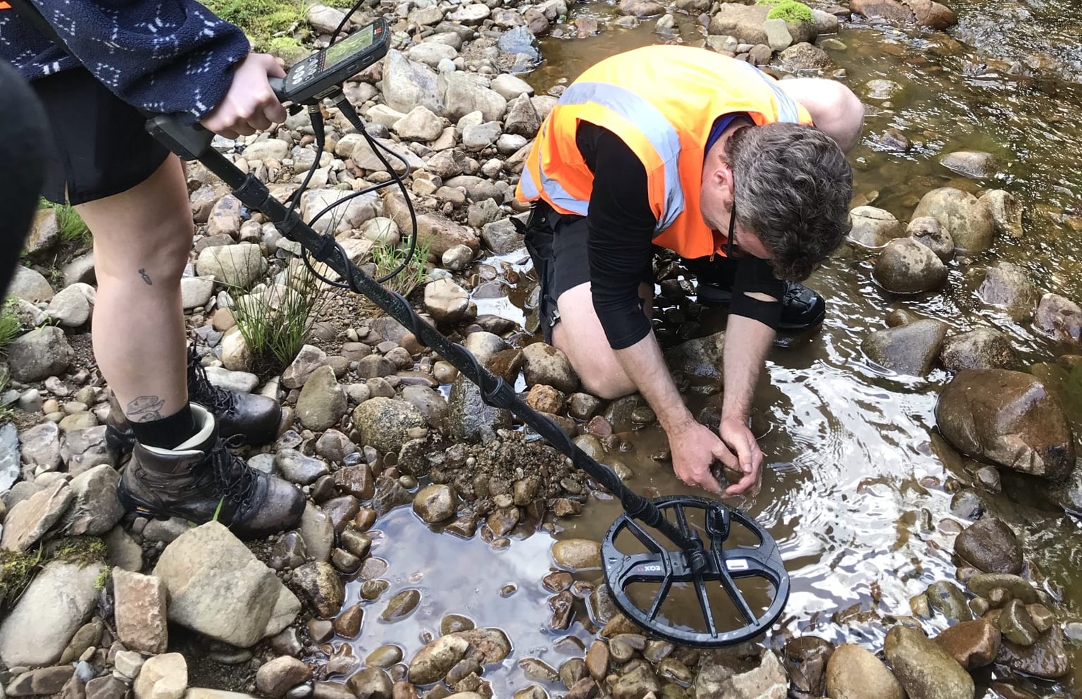 Nadine Cooper & James Scott search the riverbed to find what the metal detector has picked up. James is scooping stones out of the river bed while Nadine stands beside him holding the metal detector.
