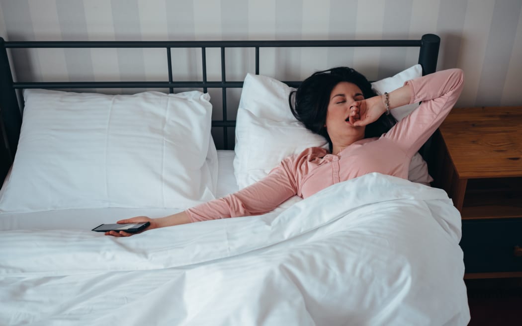 Woman waking up in her bed at home while holding a smartphone in her hand.