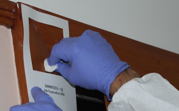A close up photo of two hands with gloves on, swabbing within a 100 square centimetre stencil stuck to a wall. By swabbing within that area, testing companies get a sample that corresponds to Ministry of Health guidelines for meth.