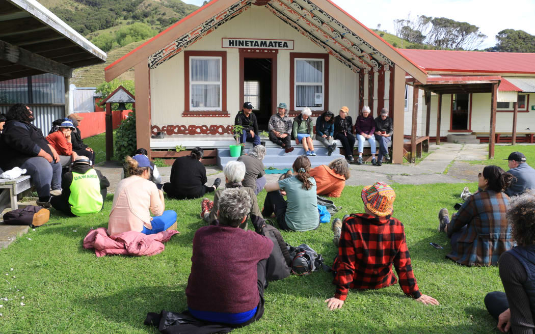 About a dozen people sitting on the lawn in front of the wharenui at Hinetamatea Marae. A further seven people are sitting on the veranda and front steps of the wharenui, which has a red roof and patterned ceiling visible under the veranda roof.