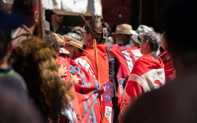 A group of wāhine Māori from Te Tai Tokerau stand on the marae atea with their backs turned on the Government in protest during Waitangi commemorations 2025. They are draped in homemade red woollen blankets hand-stitched with Tino Rangatiratanga and He Whakaputanga flags.