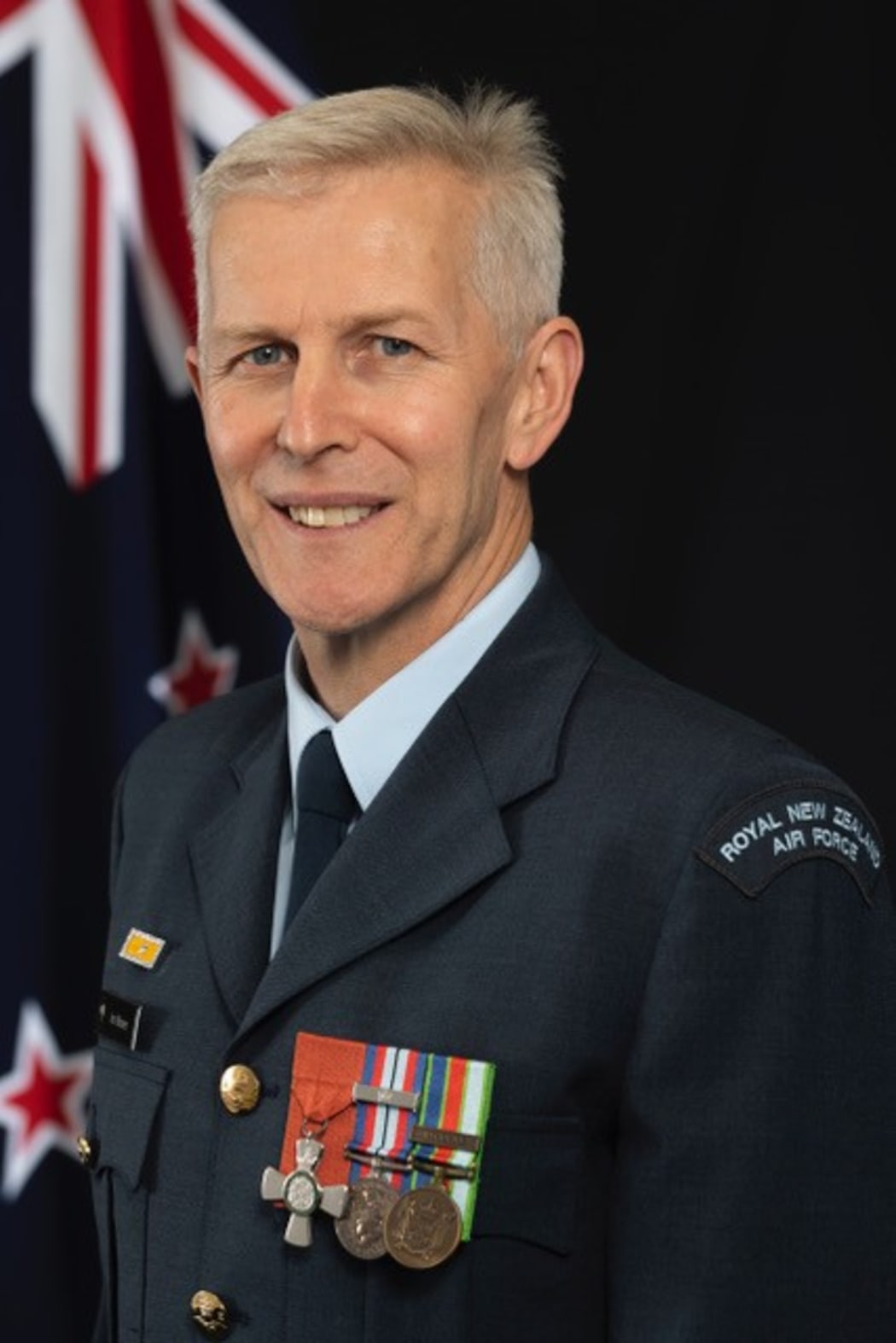 Ian smiles at the camera. He is wearing air force uniform and has badges on the left side of his chest. A New Zealand flag is hung behind him.