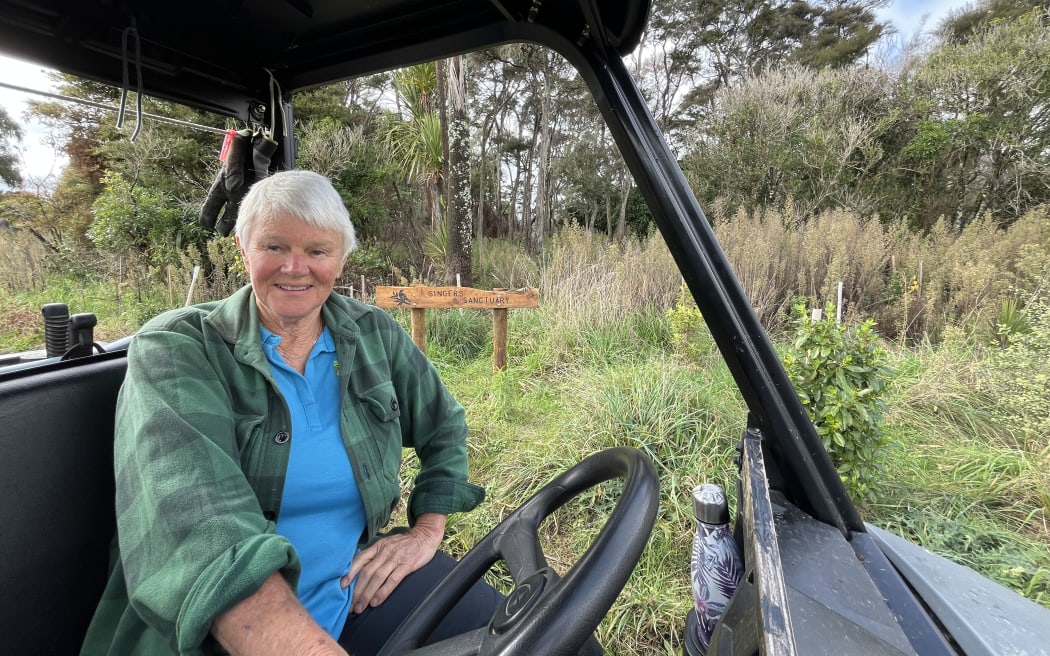 A woman in a bright blue polo shirt and a green flannel long-sleeve shirt sitting at the wheel of a buggy in front of trees and vegetation with a wooden sign. She is smiling at the camera.