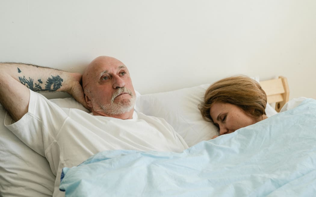 A senior couple are in bed with the woman sleeping and the man awake.