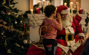 Child whispers into Santa's ear in a room with a fireplace and Christmas tree.