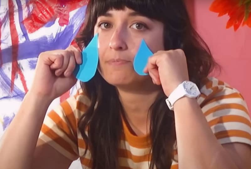 A dark haired woman in a striped t-shirt holds blue cut-out tears on her cheeks.