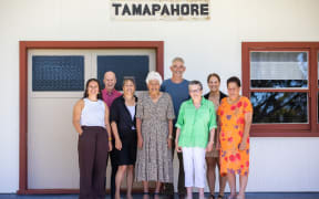 Eight people stand clustered in a group in front of a building beneath a sign that reads 'Tamapahore'. The building is white with brown-framed windows and double-door. The people are smiling at the camera for the photo.