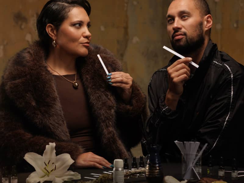 Whitney Steel & Nathan Taare sit at a table and smell samples of a perfume. White text below them reads: "JOURNEY OF SCENT."