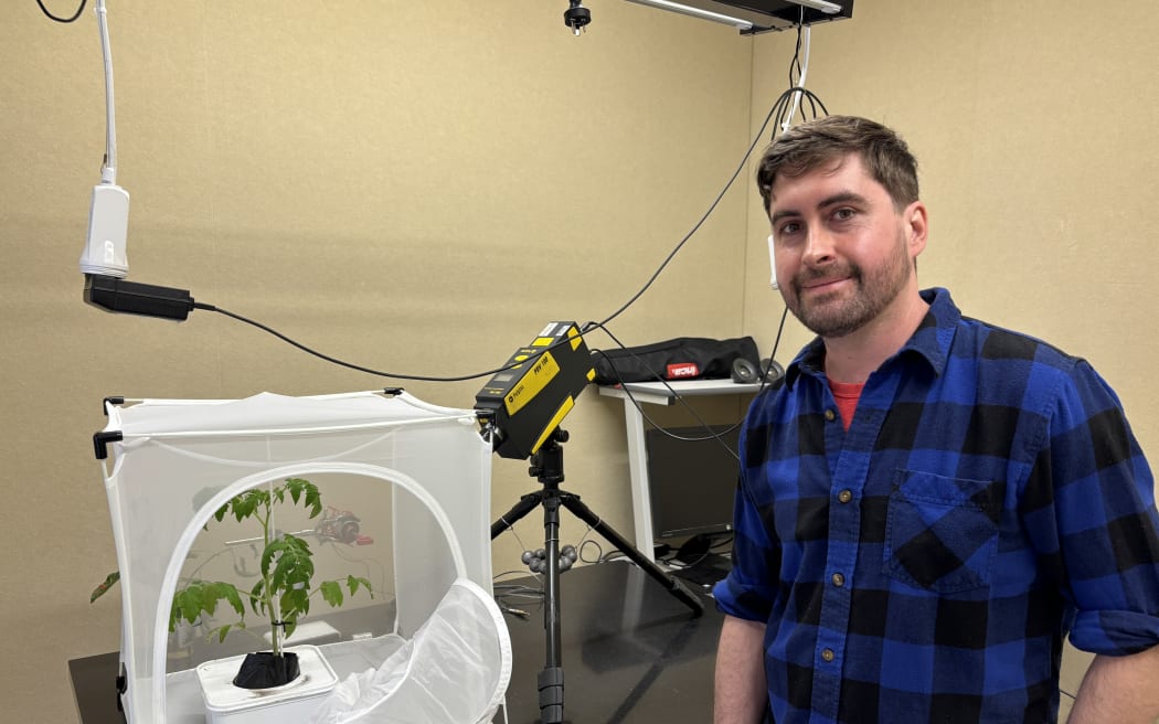 Mark stands in front of a table that has a tripod with a yellow and black laser vibrometer on top of it (a rectangle box that looks kind of like a camera). It is pointing at a small white mesh cube enclosure that has a single tomato plant inside it. Mark is smiling at the camera, and is wearing a blue and black checked shirt.