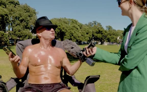 A shirtless man is interviewed by a woman in a green jacket