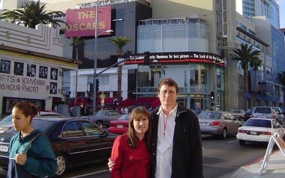 'Tania and Richard stand on a street corner smiling at the camera. Behind them is a busy Los Angeles street with cars zooming past. In the background, a large building bears a red sign reading "THE OSCARS".