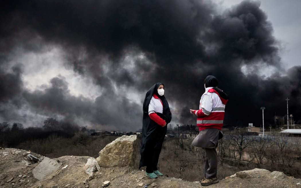 Two women from the Iranian Red Crescent Society stand as a thick plume of smoke rises in the sky in Tehran on Sunday.