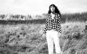 This is a black and white photo of Susan O'Neill. She is standing in a field of long grass. Susan wears a check shirt. It is impossible to say which colour because the photo is black and white. She has long, slightly wild hair. Judging by her hair it is moderately windy.