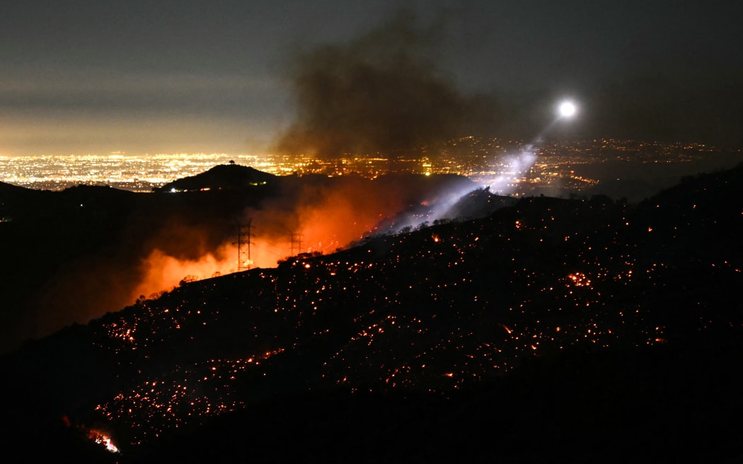 The light of a fire fighting helicopter illuminates a smouldering hillside as the Palisades fire grows near the Mandeville Canyon neighborhood and Encino, California, on January 11, 2025. The Palisades Fire, the largest of the Los Angeles fires, spread toward previously untouched neighborhoods on January 11, forcing new evacuations and dimming hopes that the disaster was coming under control. Across the city, at least 11 people have died as multiple fires have ripped through residential areas since January 7, razing thousands of homes in destruction that US President Joe Biden likened to a "war scene." (Photo by Patrick T. Fallon / AFP)