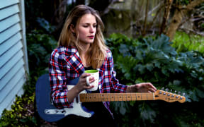 A colour photo of a young Women sitting down holding a guitar and a coffee cup