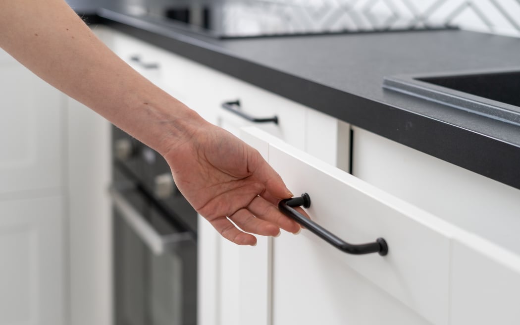 Cropped shot of female hand opening white wooden cupboard door with dark metal handle at kitchen with contemporary furniture.