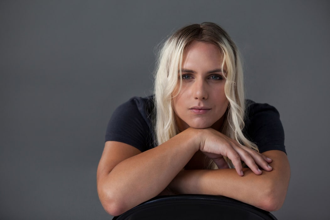 A close up photo of a blonde transgender woman resting her head on her hands and leaning forward towards the camera